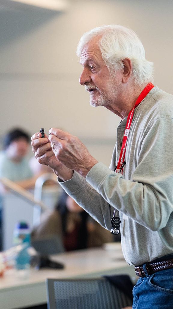 Photo: An older man in jeans and a buttoned shirt holds a rock as he teaches a class.