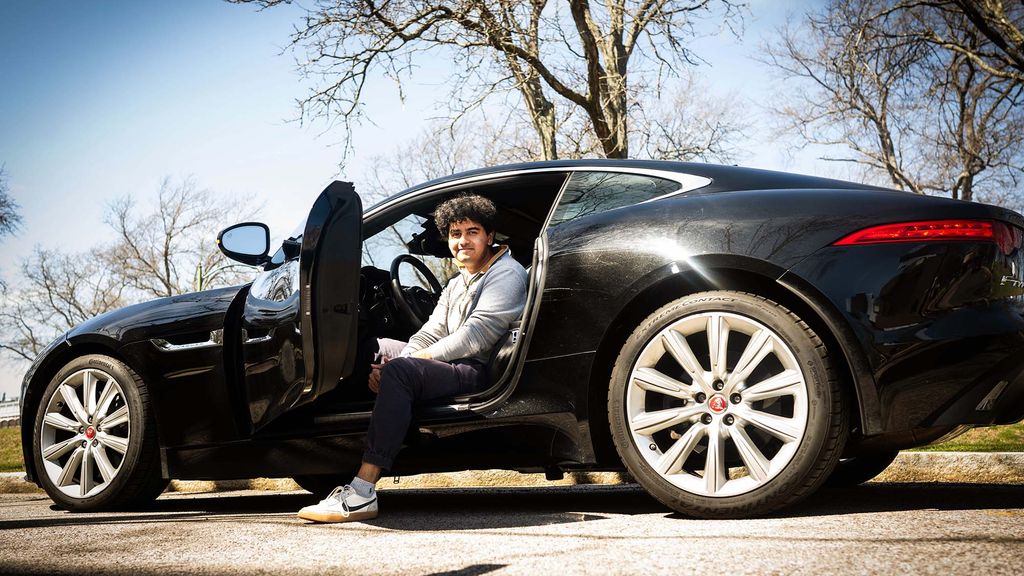 Photo: A male BU student sitting in a black sedan with the door open and one leg on the sidewalk outside of the car.