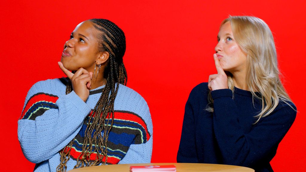 Photo: Two female BU students wearing blue sweaters posing inquisitively in front of a red background as they sit behind a table