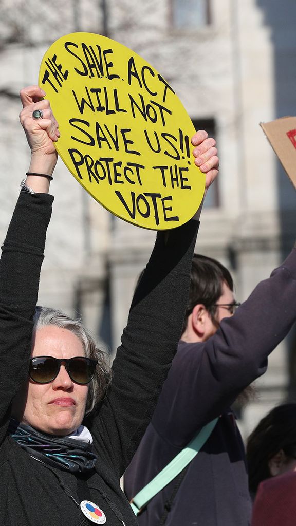 Photo: People protesting outside of a capitol building holding up signs. A woman is holding a circular sign that says "The SAVE Act Will not Save Us! Protect the Vote" and a man is holding a sign that says "People over Profits"
