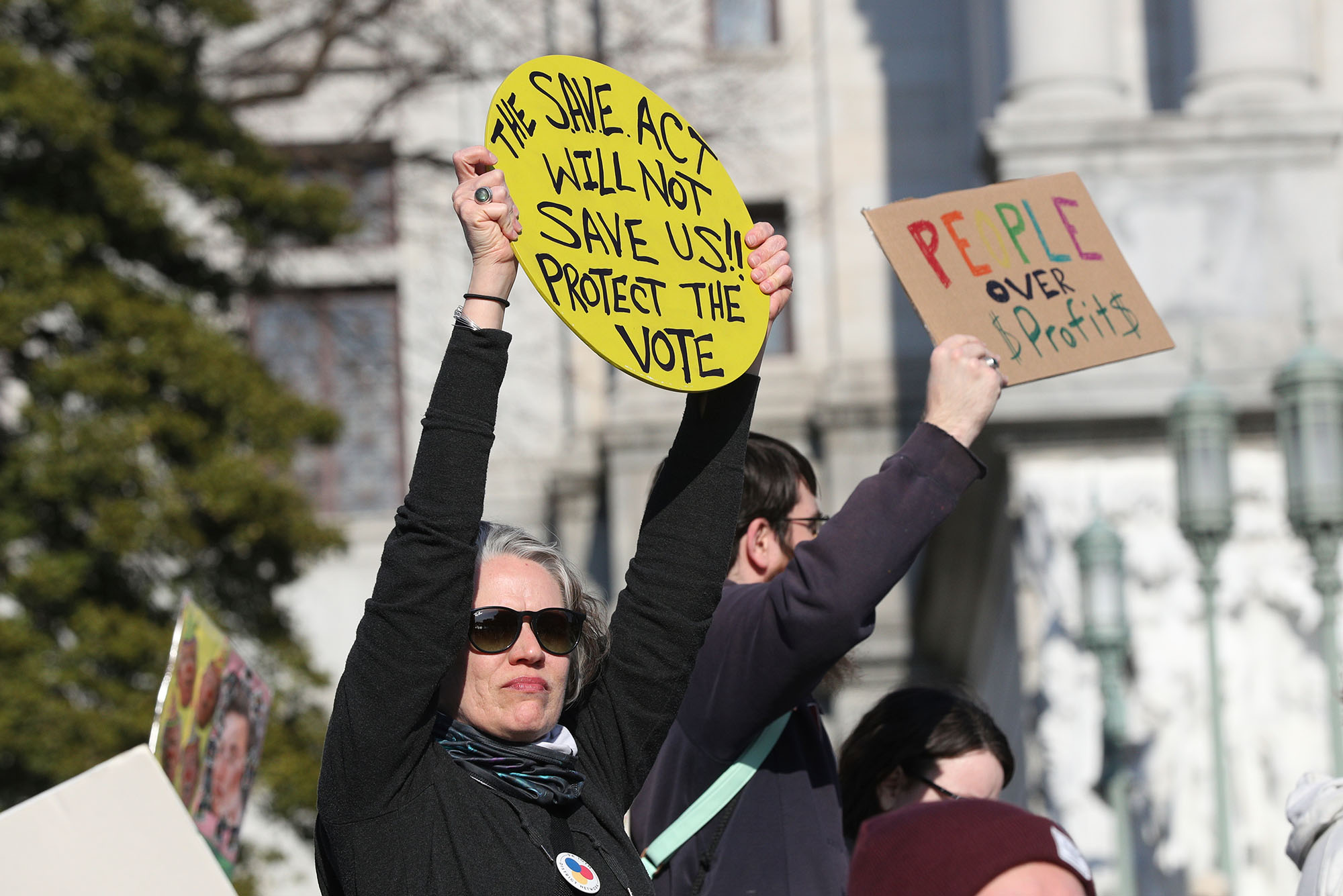 Photo: People protesting outside of a capitol building holding up signs. A woman is holding a circular sign that says "The SAVE Act Will not Save Us! Protect the Vote" and a man is holding a sign that says "People over Profits"