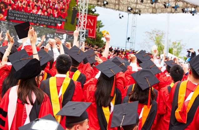Boston University students in their caps and gowns, looking up at the stage at Commencement on Nickerson Field.