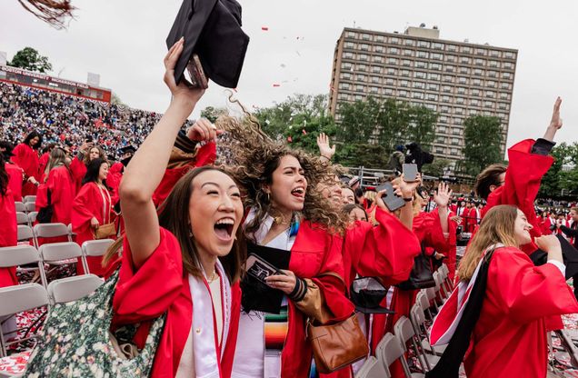 Photo: A group of Boston University graduates in red gowns and caps celebrate at an outdoor graduation ceremony. Some are raising their hands and holding diplomas, while others have their faces blurred. The background shows a large crowd of spectators in a stadium and a tall building.