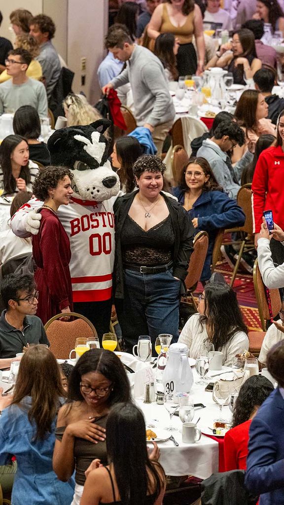 Photo: A large banquet hall filled with numerous round tables, each set with white tablecloths, plates, glasses, and cutlery. The room is crowded with people sitting at the tables and standing around. In the center of the image, a person in a Boston University mascot costume (Rhett) wearing a red and white jersey with "BOSTON 00" on it is posing for photos with attendees. Many people are taking pictures or looking at their phones. The atmosphere appears lively and social.