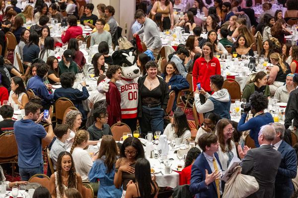 Photo: A large banquet hall filled with numerous round tables, each set with white tablecloths, plates, glasses, and cutlery. The room is crowded with people sitting at the tables and standing around. In the center of the image, a person in a Boston University mascot costume (Rhett) wearing a red and white jersey with "BOSTON 00" on it is posing for photos with attendees. Many people are taking pictures or looking at their phones. The atmosphere appears lively and social.